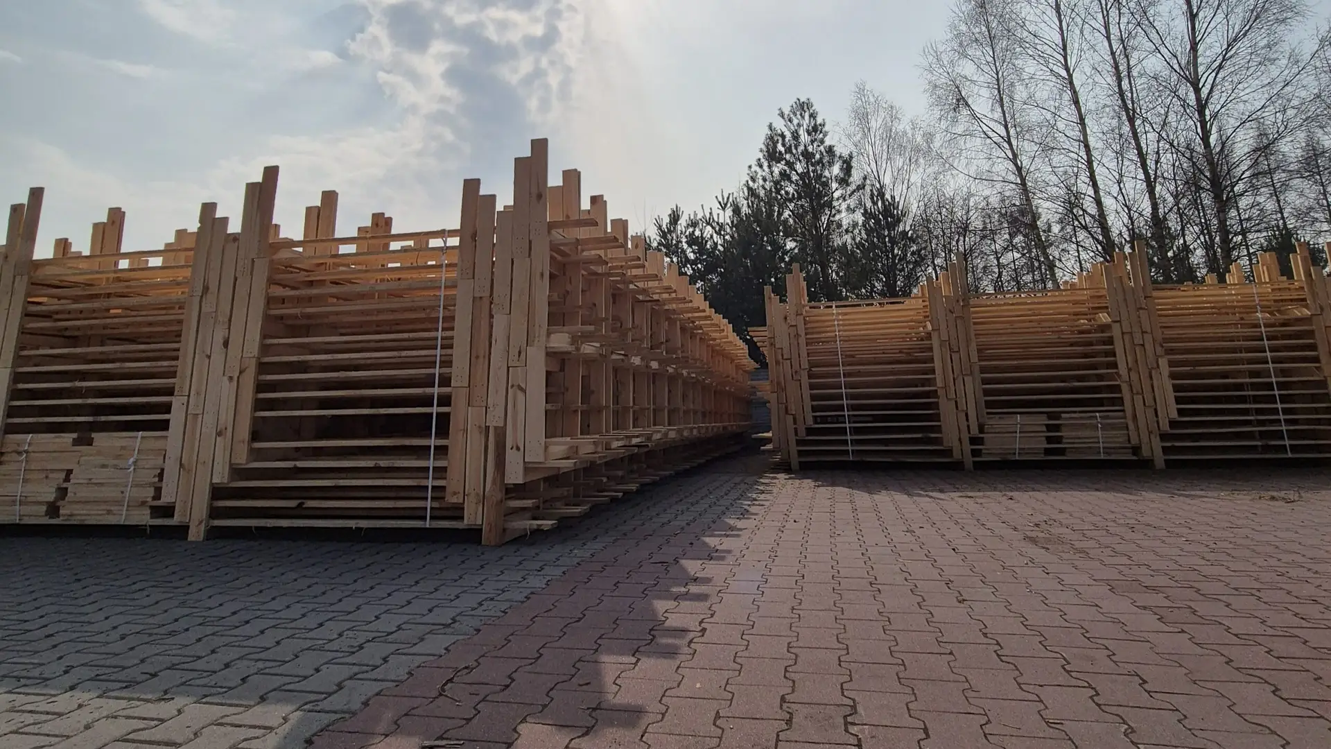 Stosy zbitek drewnianych starannie ułożonych na wybrukowanej ścieżce pod zachmurzonym niebem, z drzewami w tle i przebijającymi się przez nie promieniami słońca.