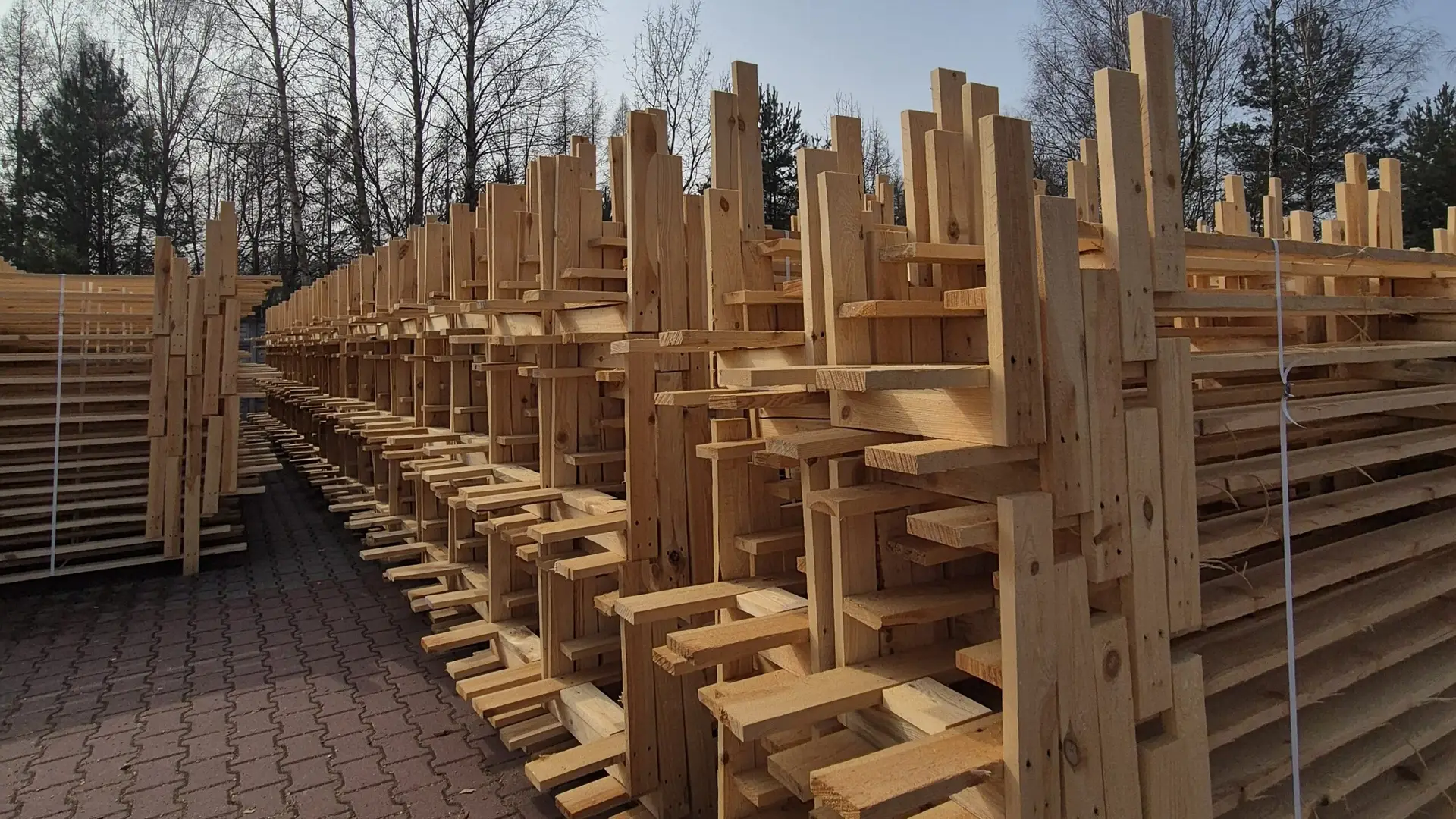 Długi stos starannie ułożonych zbitek drewnianych wzdłuż ceglanej ścieżki pod czystym niebem, z drzewami w tle.