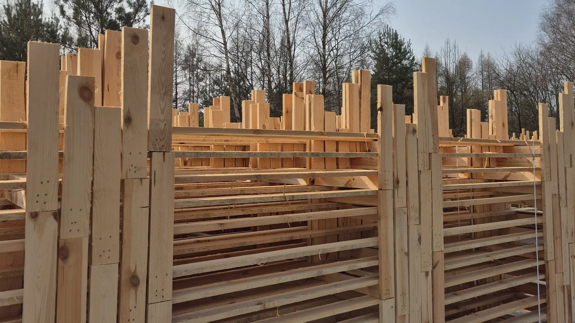 Stosy zbitek drewnianych starannie ułożone na zewnątrz, z drzewami i czystym, błękitnym niebem w tle.
